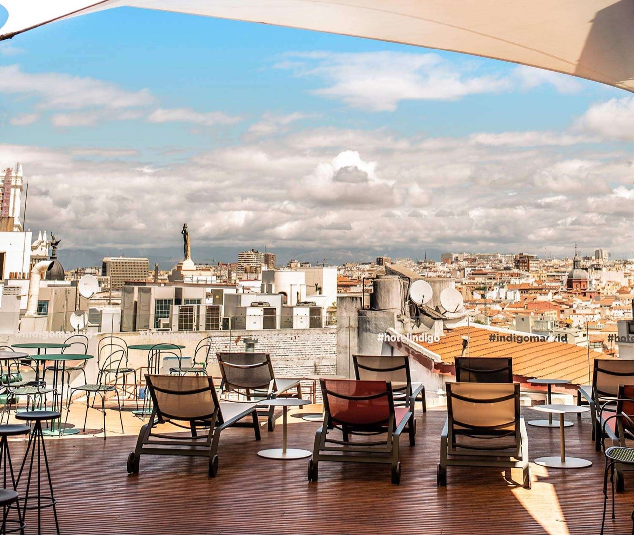 Rooftop terrace with wooden decking, loungers and small tables under an awning, overlooking Madrid's rooftops with '#hotelindigo' printed on the awning.