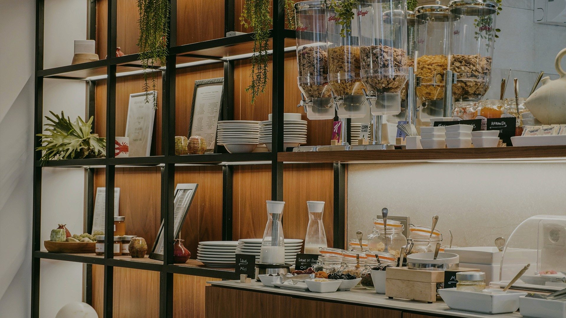 Breakfast buffet counter at Hotel L'Avenida with cereal dispensers, stacked white plates, and hanging plants against a wood-shelved wall