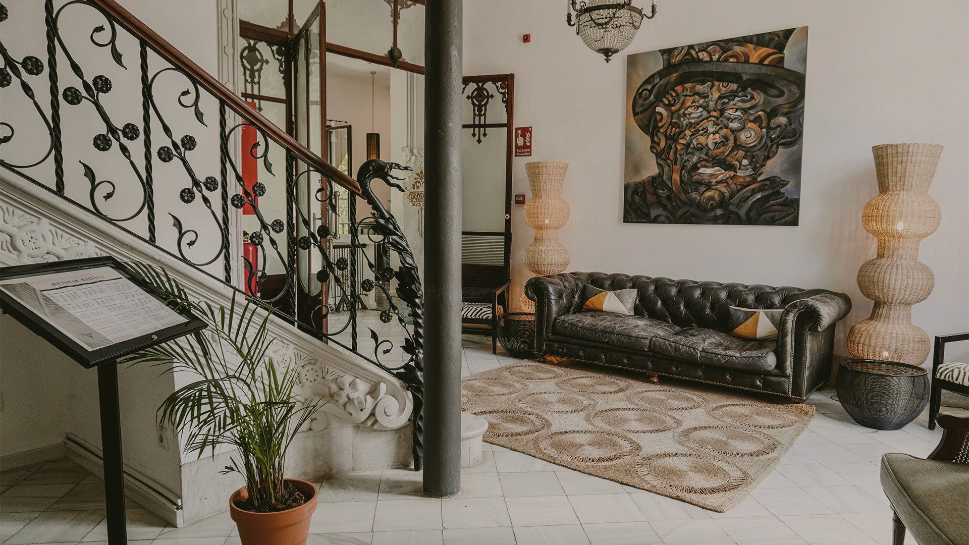 Lobby at Hotel L'Avenida with an ornate wrought-iron staircase, a black leather chesterfield sofa, and a large modern painting on the wall