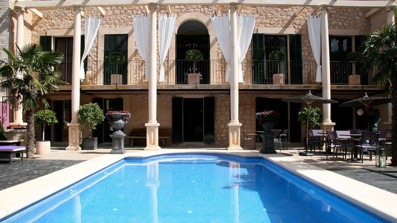 Courtyard swimming pool at Hotel L'Avenida framed by a stone colonnade, draped white curtains, and the historic building's shuttered balconies
