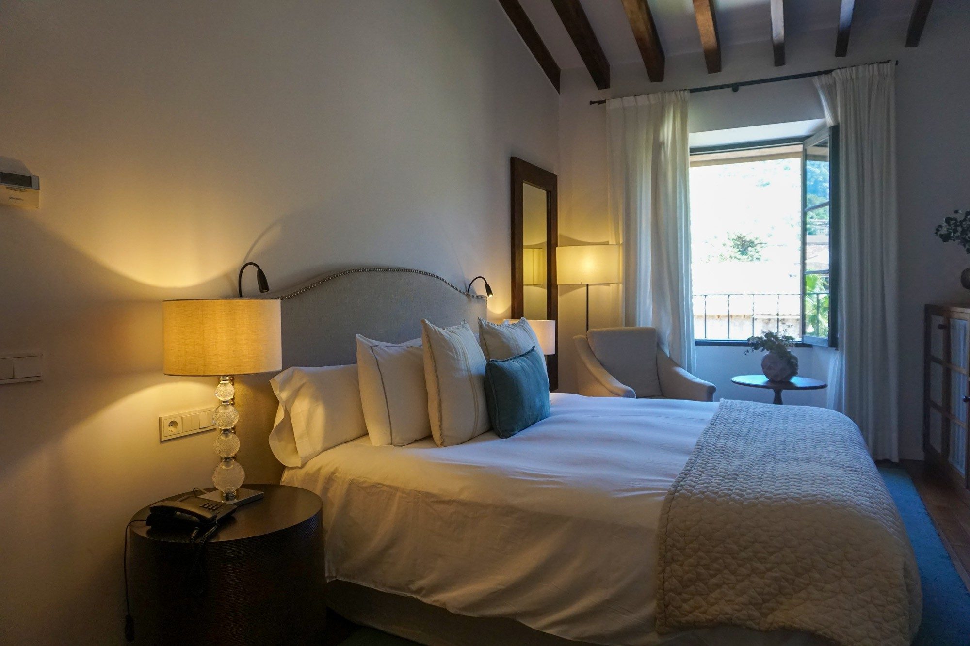 Double room at Hotel L'Avenida with a linen headboard, teal accent pillows, and an exposed wood-beam ceiling above a window onto Soller's old town