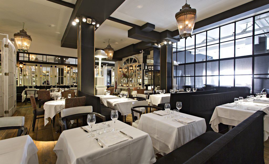 Market Restaurant dining room with white-clothed tables, dark banquette seating, wrought-iron lantern pendants and tall black-framed windows