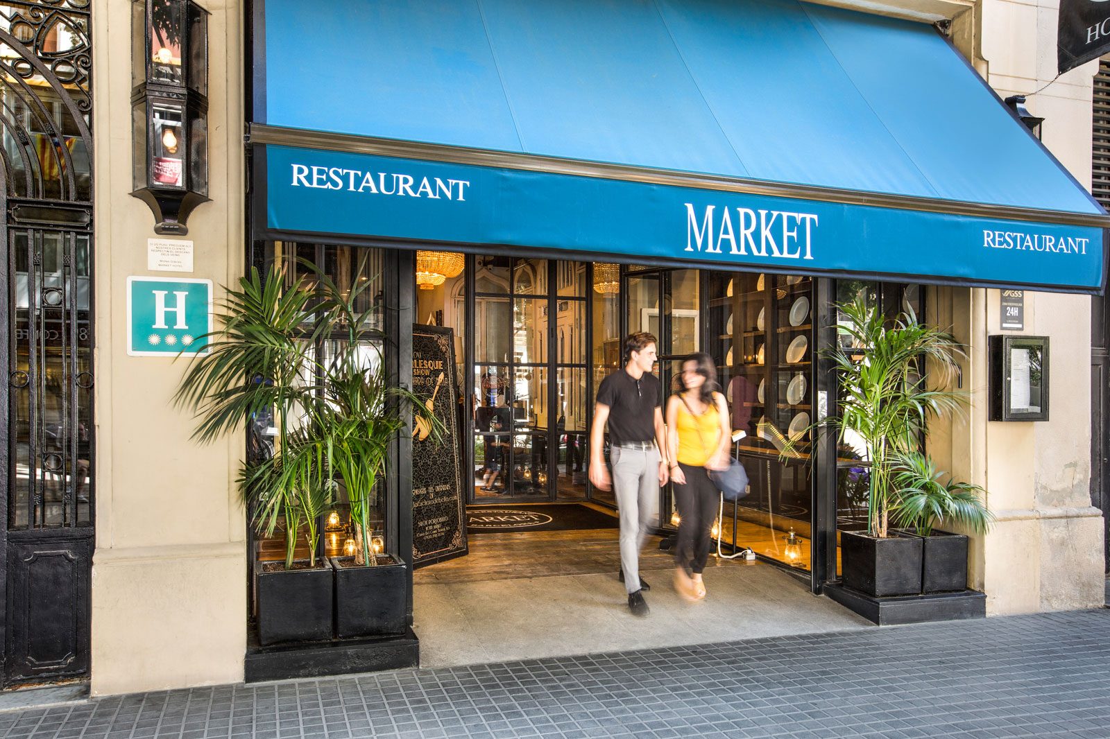 Street-level entrance of Hotel Market under a blue Market Restaurant awning, with black wrought-iron doors and potted palms flanking the doorway