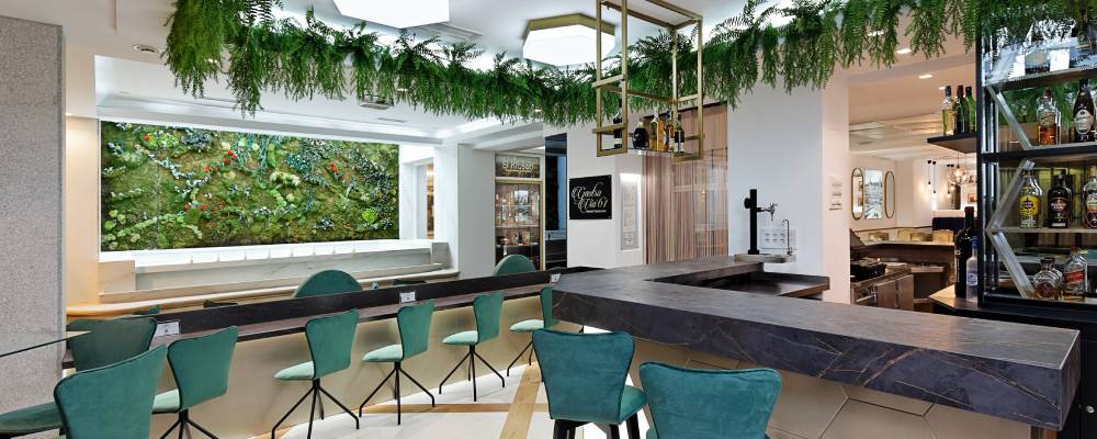 Bar Retiro with teal upholstered stools along a marble-topped counter, a large vertical garden panel of preserved plants on the wall and hanging greenery from the ceiling.