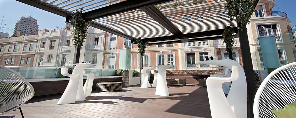 Rooftop terrace under a slatted pergola with white cocoon-style chairs and tall stool tables, overlooking neighbouring Madrid building facades.