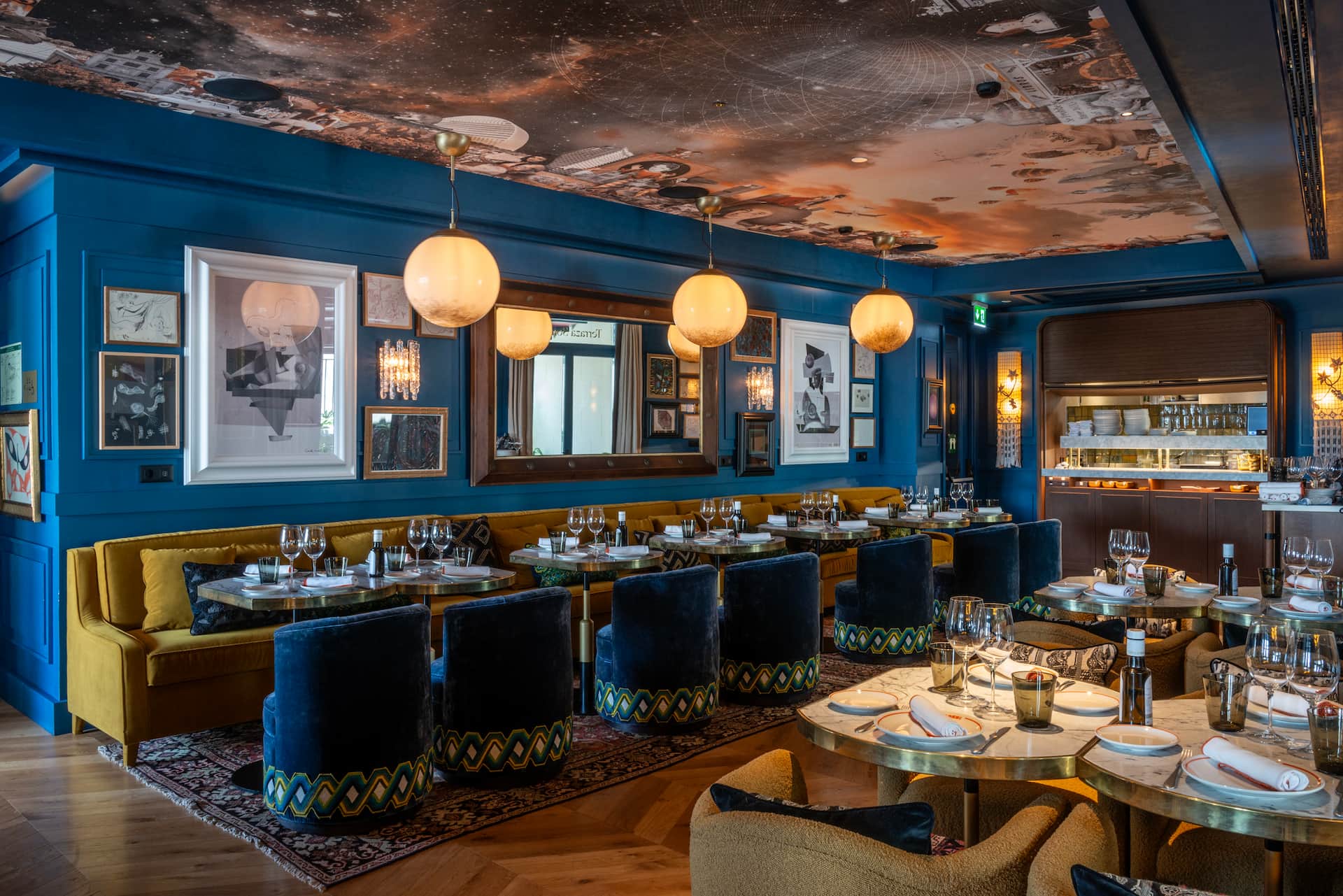 The restaurant: a blue-panelled dining room with a mustard velvet banquette, navy velvet tub chairs, brass-edged marble tables, framed artworks, globe pendants and a collage-printed ceiling.