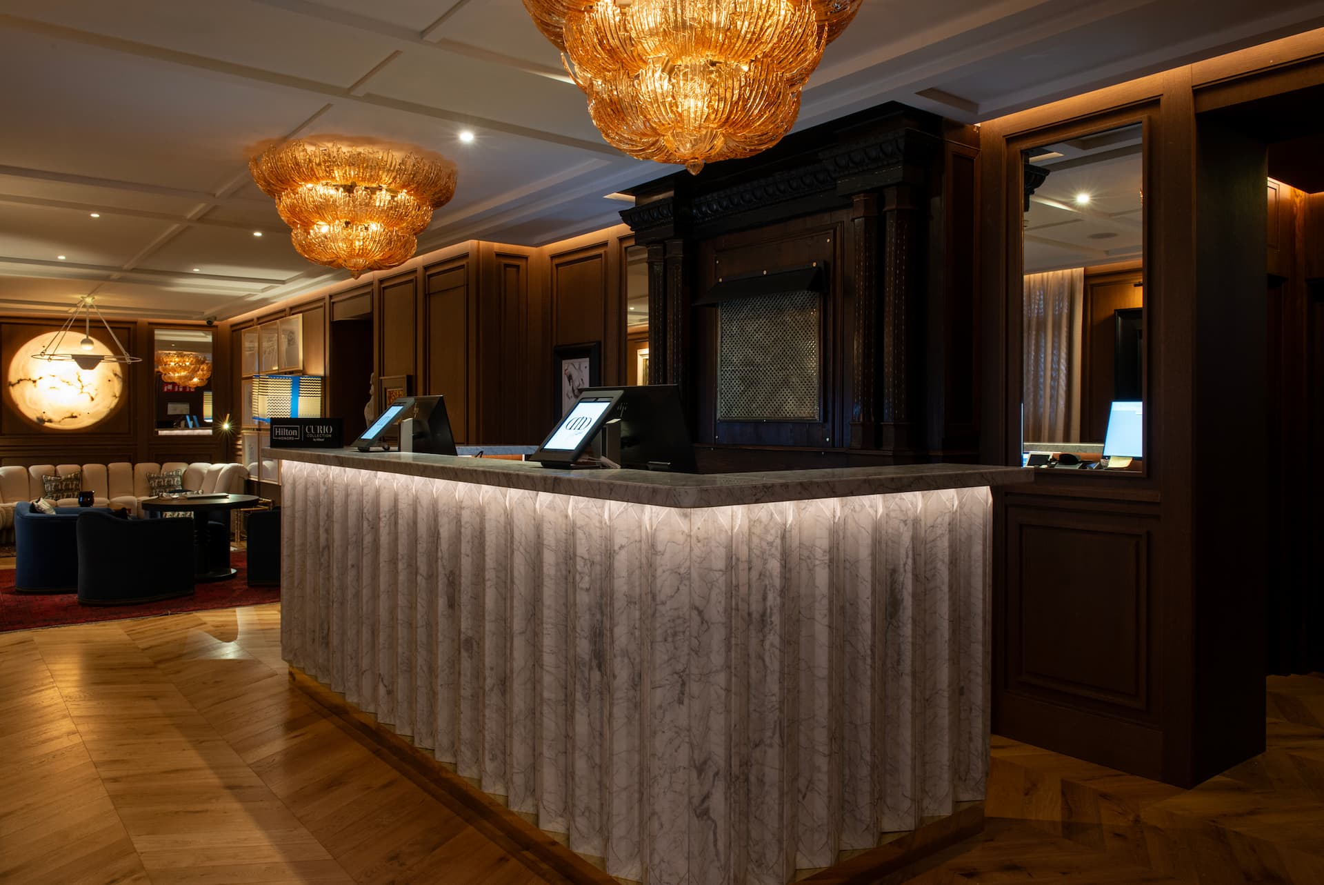 The reception: a fluted white-marble front desk lit from below, two amber glass chandeliers, dark wood panelling and a lounge with cream sofas and blue armchairs beyond.