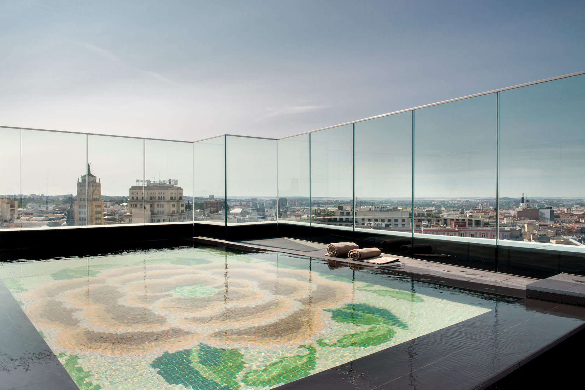 The rooftop pool: a shallow pool with a rose mosaic on its floor, rolled towels at the edge and glass railings over the Madrid skyline, the Telefonica building in the distance.