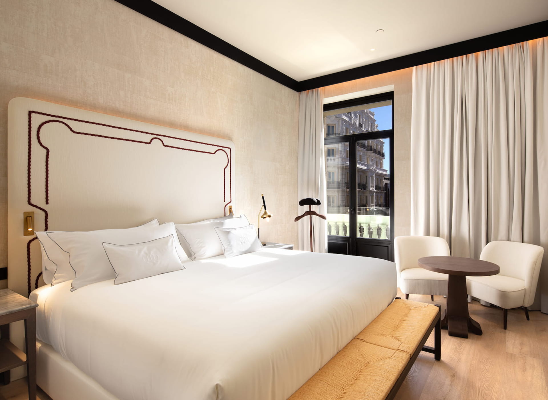 A guest room at Hotel Montera: a white bed under a cream headboard with dark-red braid trim, two cream armchairs at a round table, and a black-framed balcony door onto an ornate stone facade opposite.