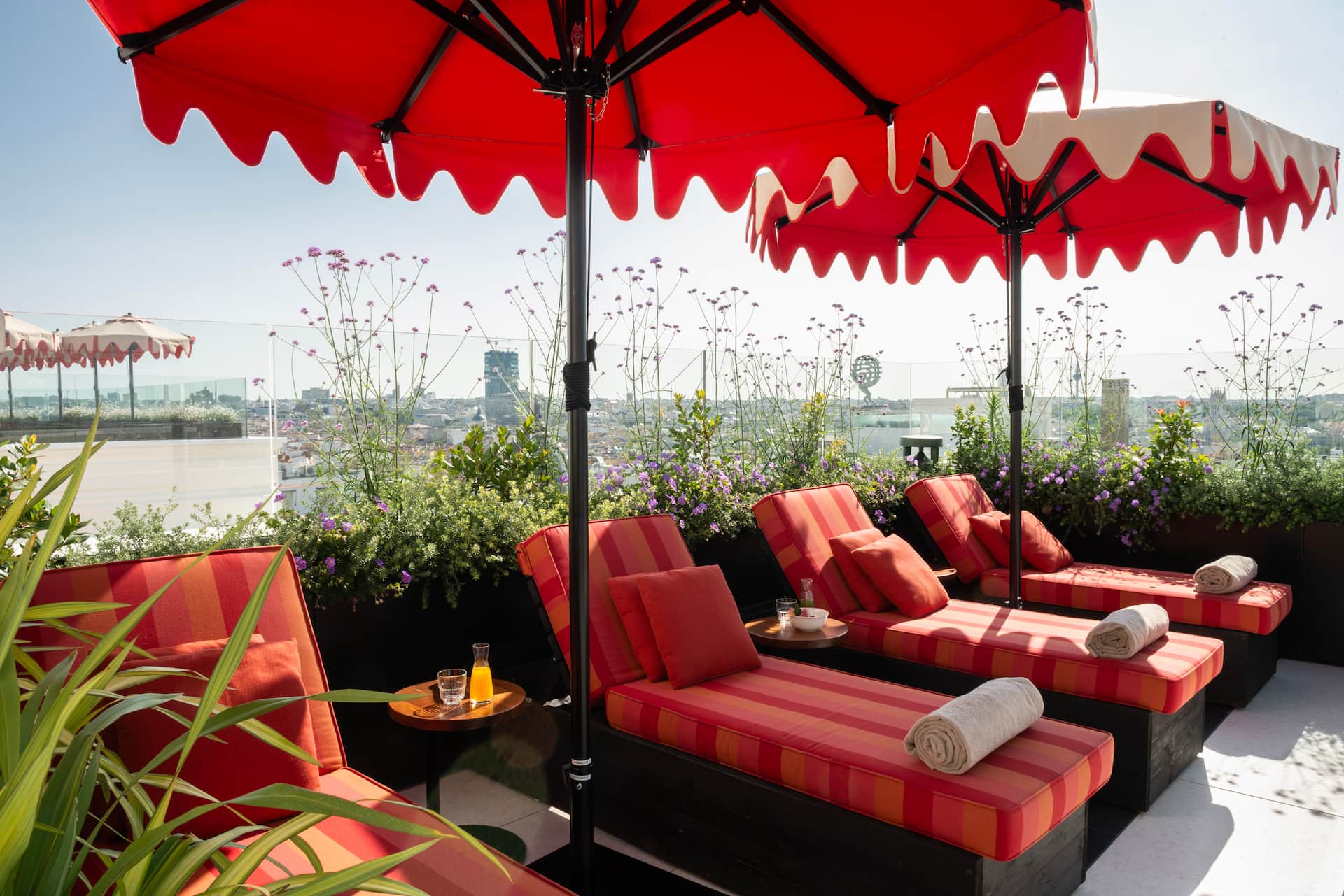 The rooftop terrace: red-and-orange striped sun loungers with rolled towels under red scalloped parasols, planters of purple flowers and the city skyline behind glass railings.