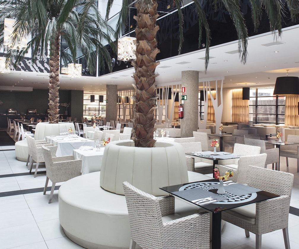 Restaurant dining room set beneath a glass roof with tall natural palm trees rising between white-clothed tables and wicker chairs.