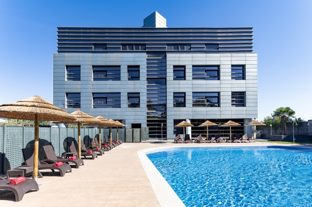 Multi-storey grey hotel building with a metal-panel facade, seen from beside the outdoor pool with thatched umbrellas and loungers in the foreground.