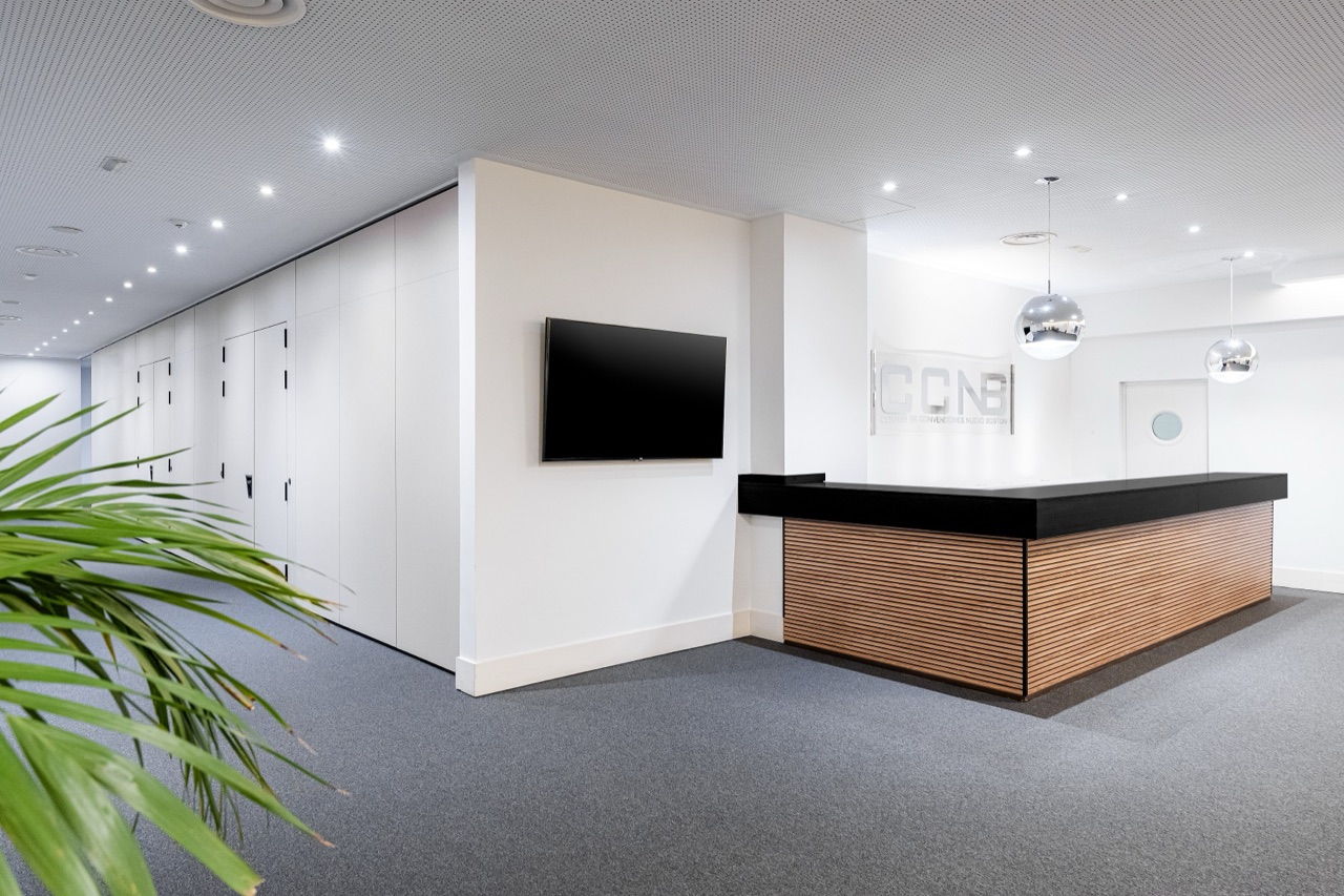 Modern reception desk in dark wood and black stone beneath a metallic 'CCNB' convention centre sign, with a wall-mounted TV and a potted palm in the foreground.