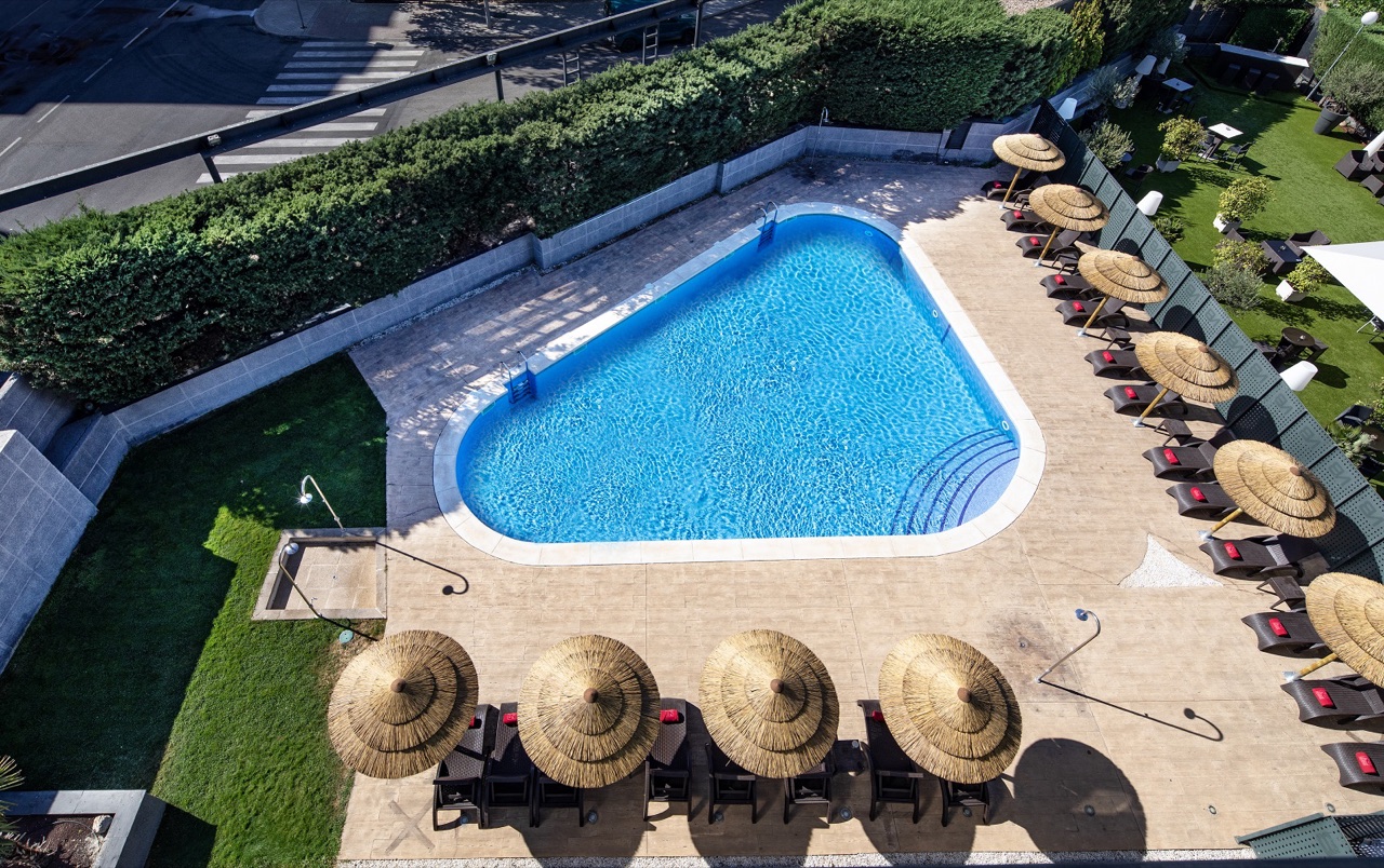Aerial view of an outdoor swimming pool lined with thatched umbrellas and lounge chairs beside the hotel building and a garden lawn.