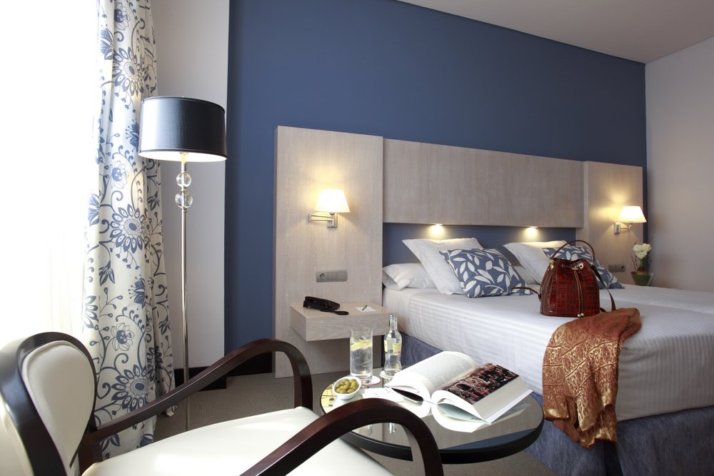 Hotel bedroom with a dark blue accent wall, wood headboard, a made double bed and a dark wood armchair beside a floral-curtained window.