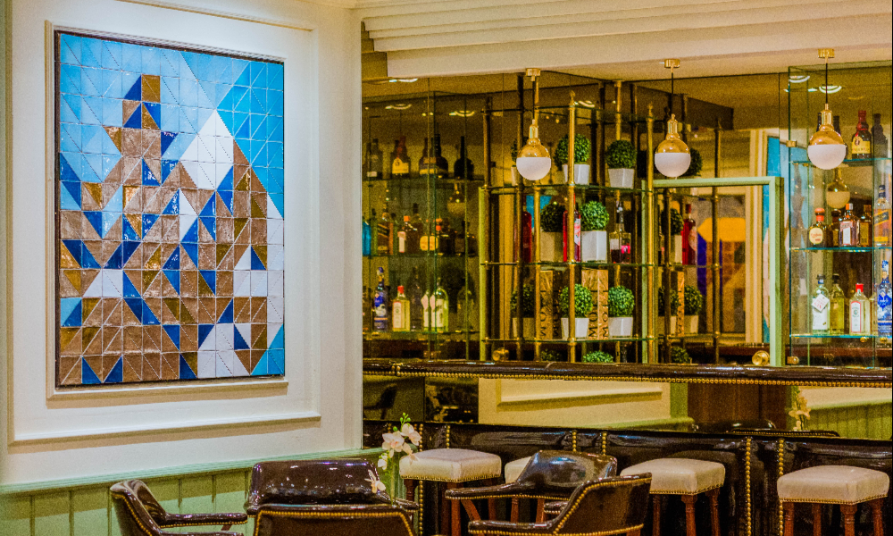 A hotel bar with a mirrored back-bar lined with bottles, hanging glass pendant lights, potted plants on brass shelving, and leather stools along the counter.