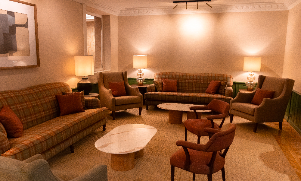 A sitting area with two plaid sofas, wingback armchairs, rust-colored cushions and table lamps arranged around two round marble coffee tables.