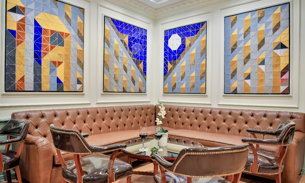 A corner lounge seating area with a tufted brown leather banquette, leather armchairs, a low table with two glasses of wine, and four geometric tiled art panels on the wall above.