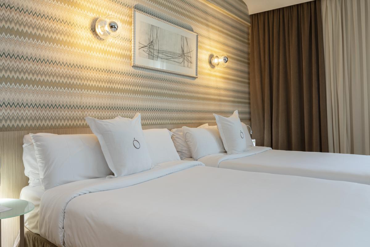 Two beds dressed in white linen against a gold and green chevron-patterned wall, with a framed abstract sketch and wall sconces above the headboard.