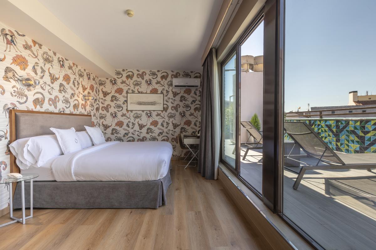 A hotel room with a bed against a zodiac-print wallpaper wall, with a glass sliding door open onto a private terrace with sun loungers and rooftop views.