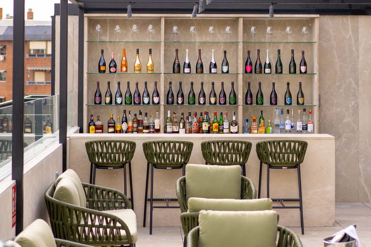 Rooftop Lounge Bar shelving stocked with wine and spirit bottles behind a stone counter, with green woven bar stools and armchairs.