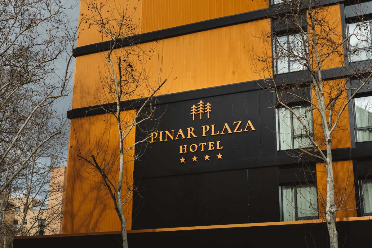 Hotel facade detail with orange and black cladding and an illuminated Pinar Plaza Hotel sign showing four stars, framed by bare trees.
