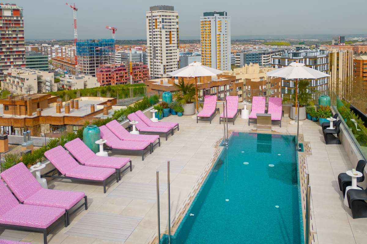Rooftop swimming pool lined with pink patterned loungers and white umbrellas, overlooking Madrid's skyline of apartment towers.