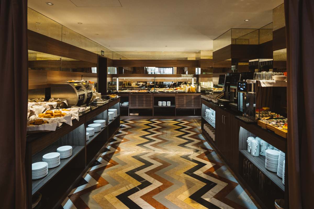 Breakfast buffet room with a chevron-patterned marble floor and dark wood counters holding pastries and stacked plates.