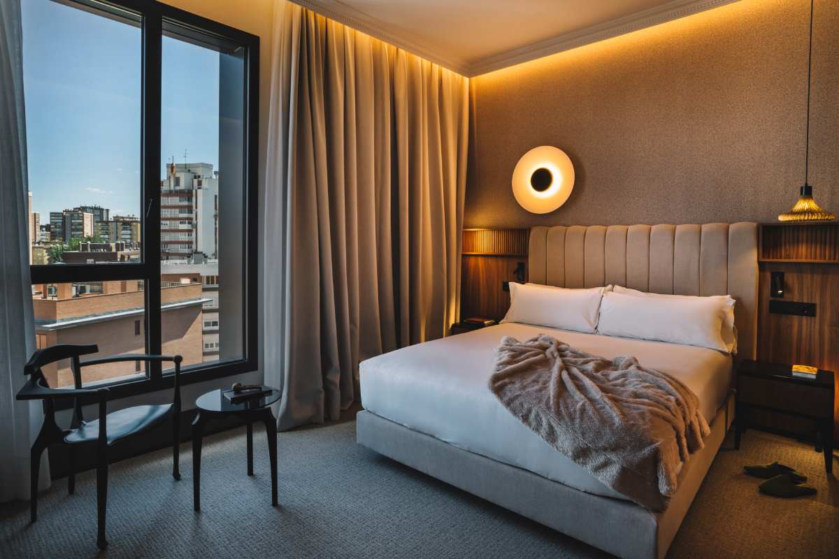 Hotel bedroom at dusk with a beige upholstered headboard, white bedding draped with a throw, and a floor-to-ceiling window over the city.