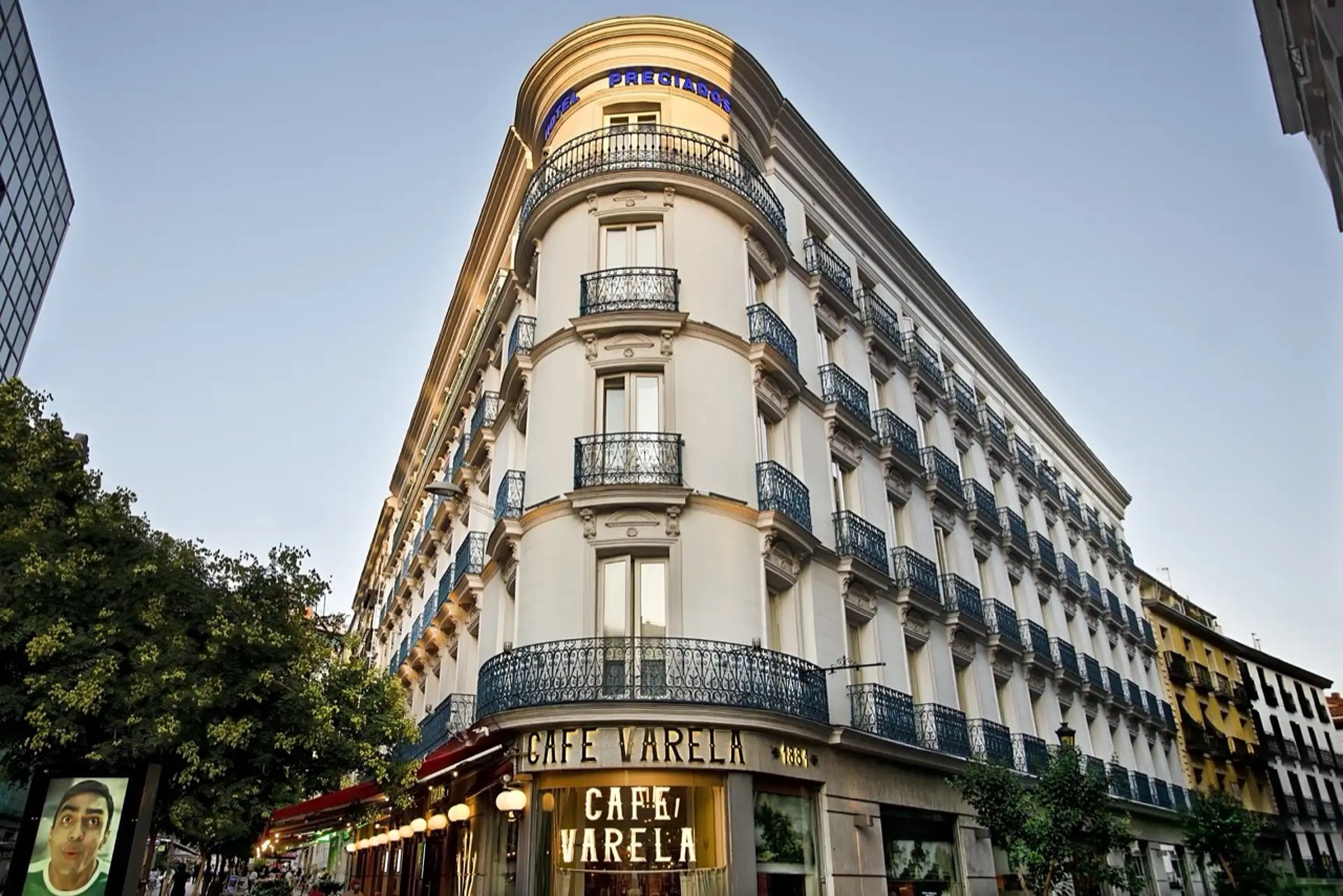Corner facade of the restored 19th-century building with wrought-iron balconies, a lit HOTEL PRECIADOS sign at the top and a CAFE VARELA sign over the ground-floor entrance.