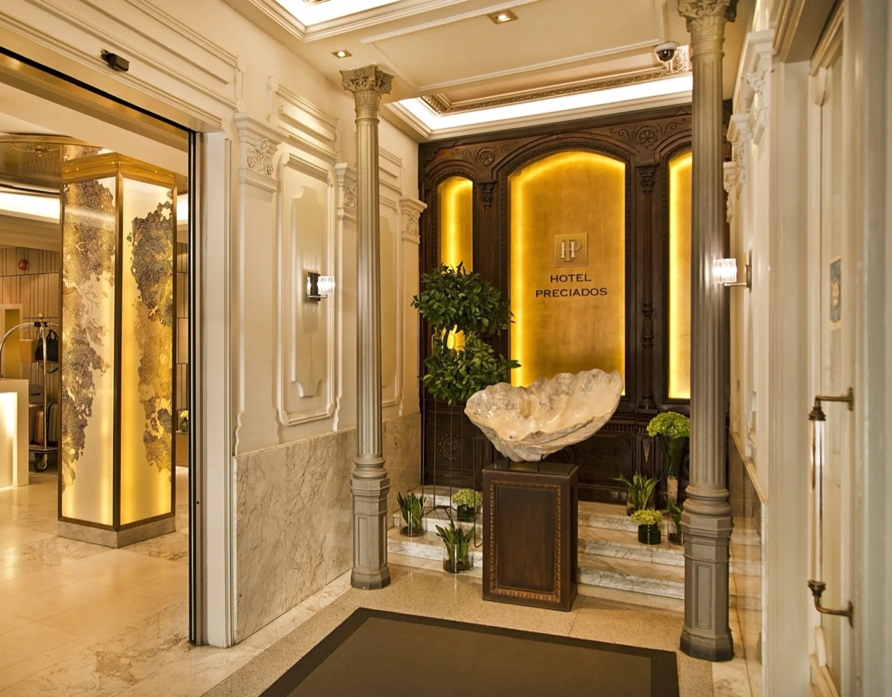 Hotel entrance hall with marble columns, a gold-lit dark wood reception wall reading HOTEL PRECIADOS and a large shell-shaped decorative basin on a pedestal.
