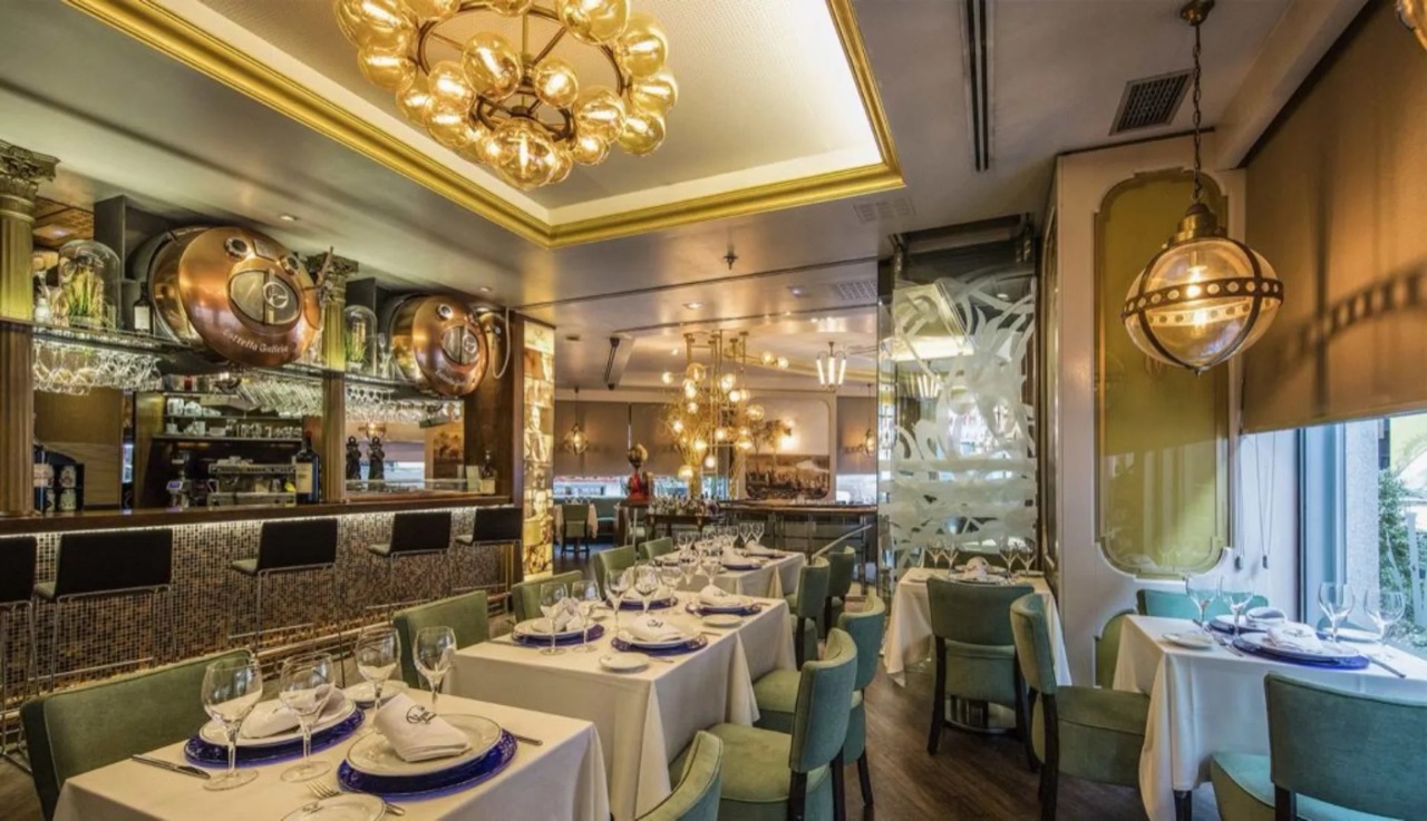Cafe Varela dining room with green upholstered chairs at white-clothed tables, a gold globe chandelier, a mosaic-tiled bar with copper barrel taps and mirrored walls.