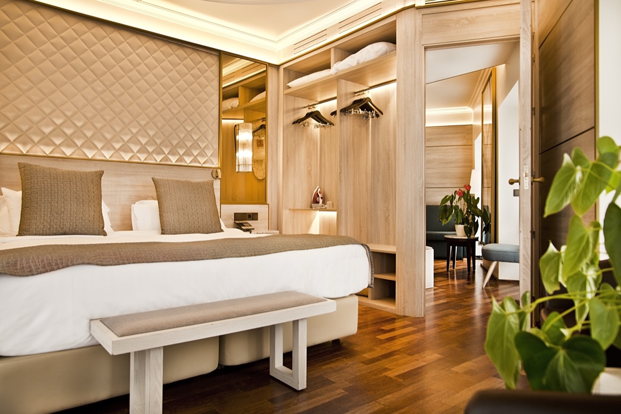 Hotel bedroom with a quilted beige headboard, a king bed with a taupe throw, a wardrobe area with hanging clothes and a wood-panelled sitting area visible through an open door.