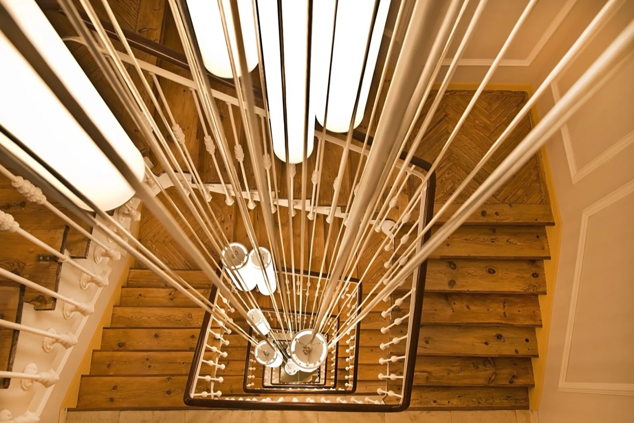 The hotel's stairwell seen from above: worn wooden stairs and a white banister winding down around a column of long pendant lights.