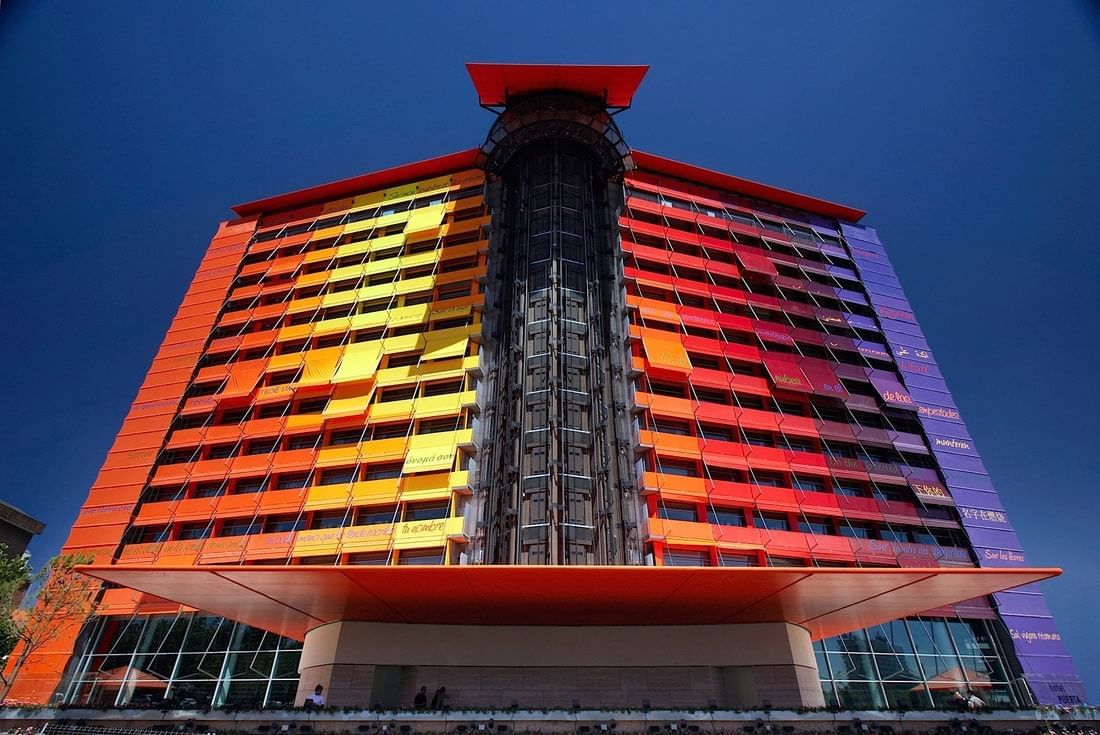 The colourful glass facade of Hotel Puerta America, shifting from yellow to red to purple across its floors, against a blue sky.