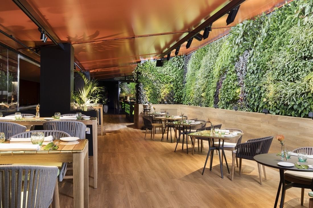 The Observatory's dining terrace with wooden tables set for service beside a large vertical garden wall under an orange awning.