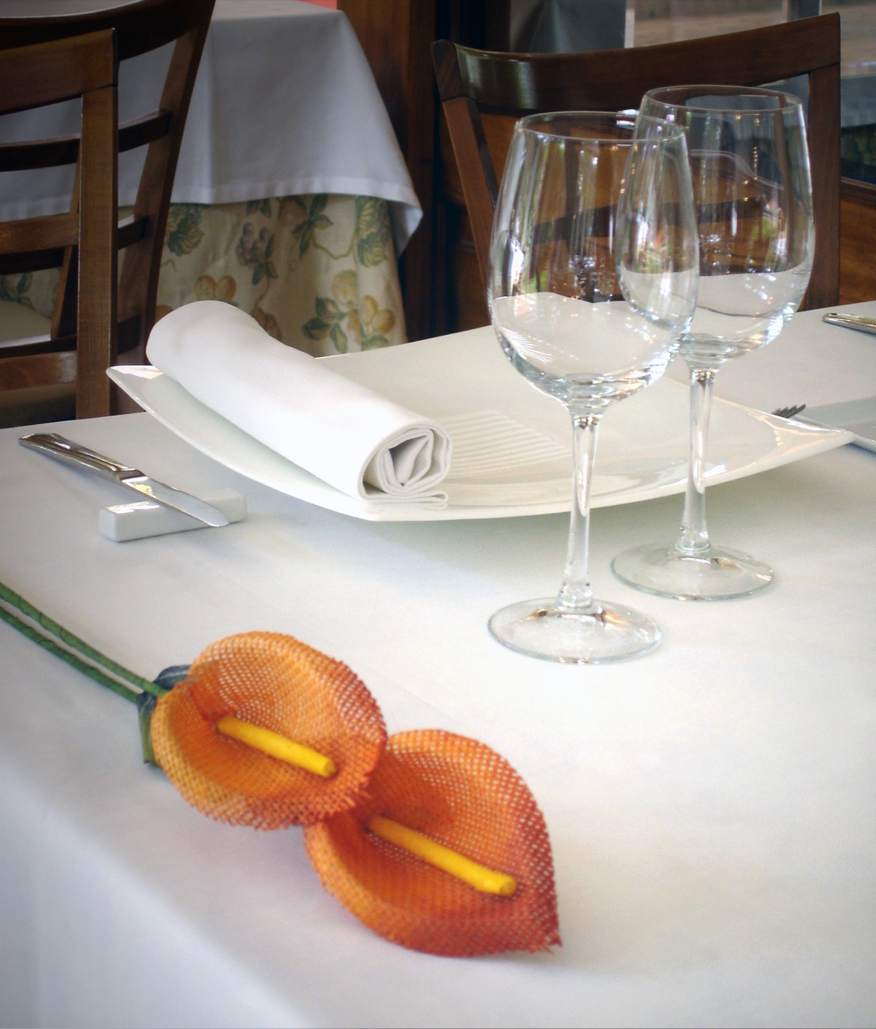 Indoor restaurant table set with white linen, wine glasses and folded napkins, wooden chairs and a floral curtain in the background.