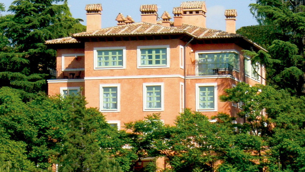 Terracotta-colored mansion building with tiled roofs and multiple chimneys, seen through surrounding cedar and pine trees.