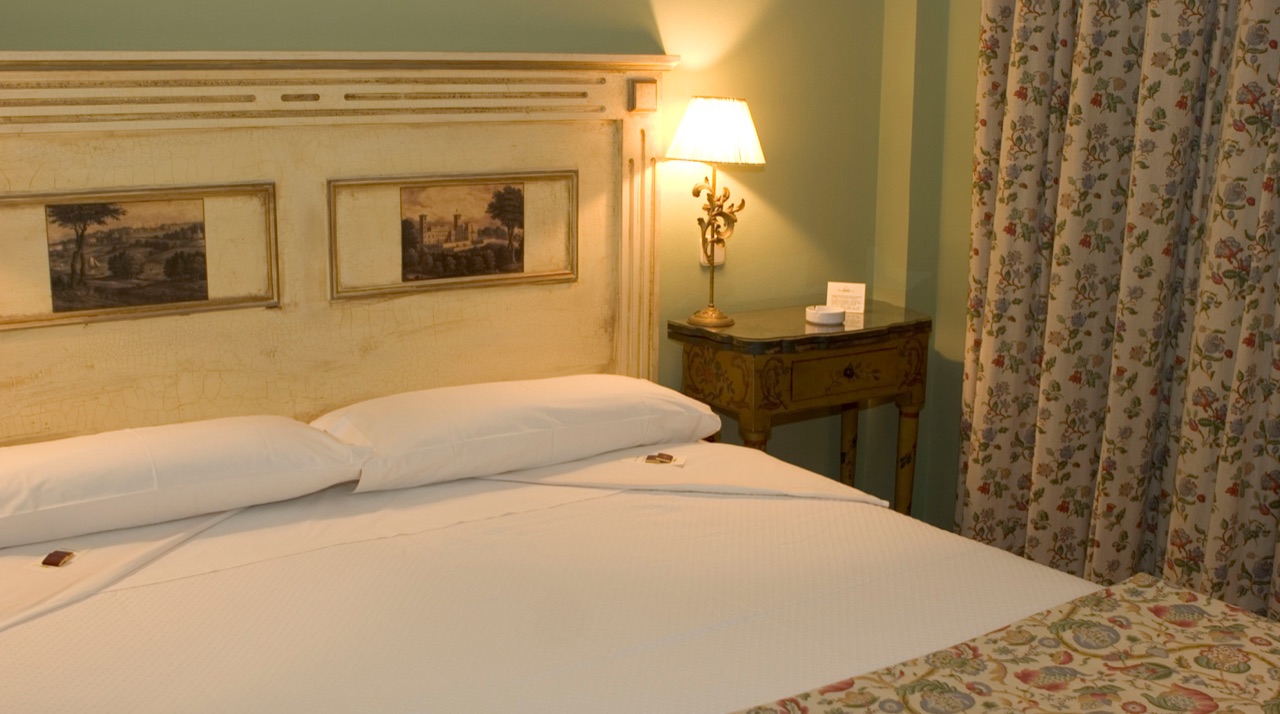 Hotel bedroom with a cream headboard holding two framed landscape prints, a bedside lamp and floral curtains.