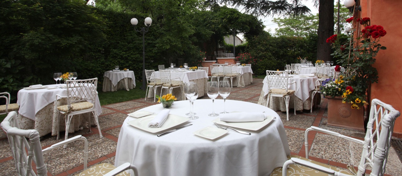 Outdoor garden terrace with white-clothed round tables and chairs set for dining among trees, flowering shrubs and a rose-covered wall.