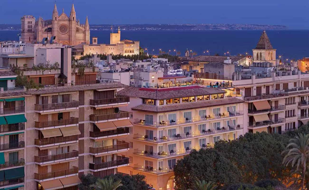 Hotel Saratoga's illuminated facade at dusk, with Palma Cathedral and the bay visible behind it.