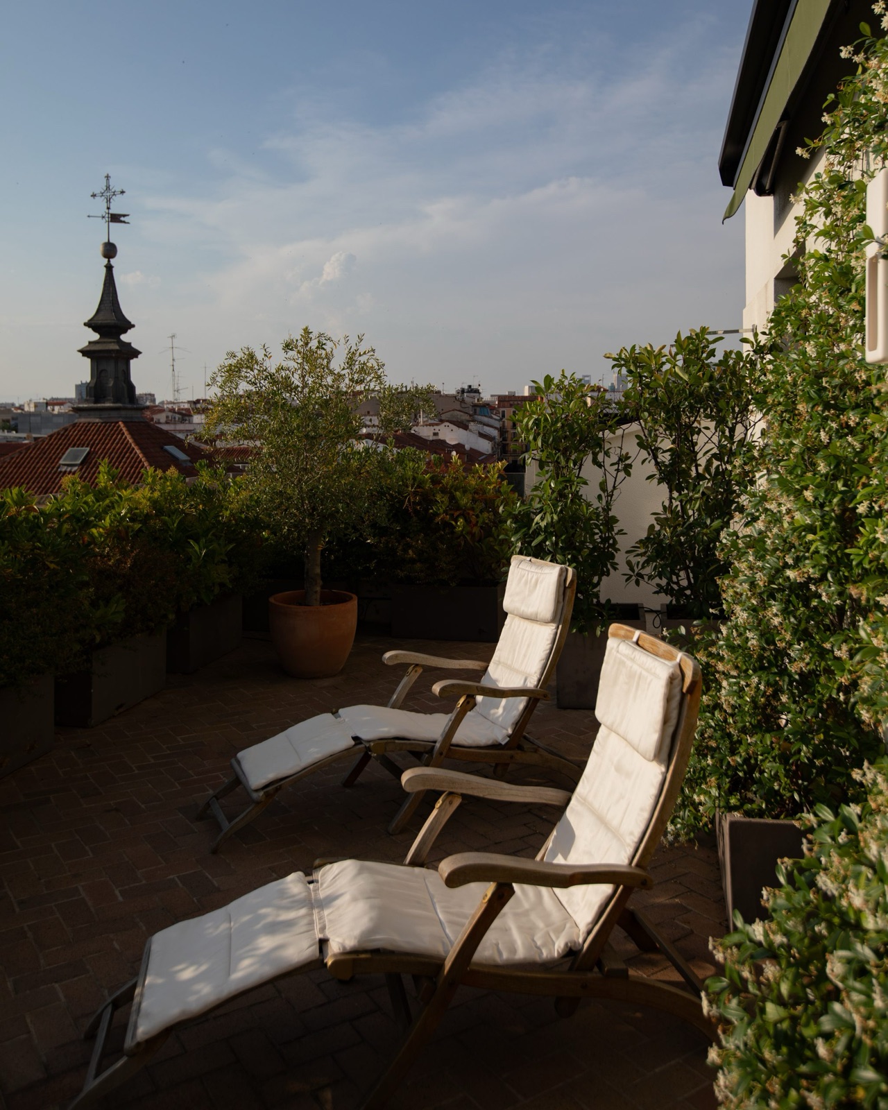 A rooftop terrace with two reclining lounge chairs, potted plants, and a view over Madrid rooftops to a church spire.