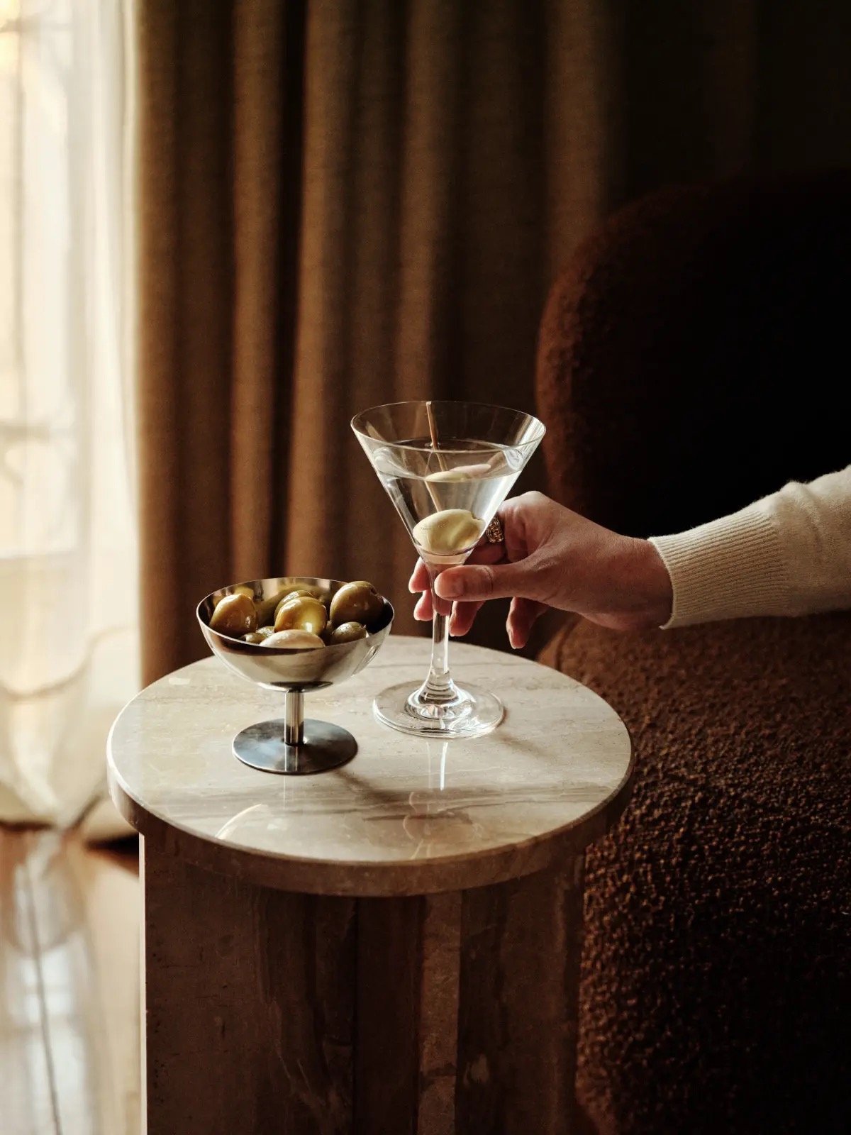 A martini with an olive and a small steel bowl of olives on a round marble side table beside a brown boucle armchair.