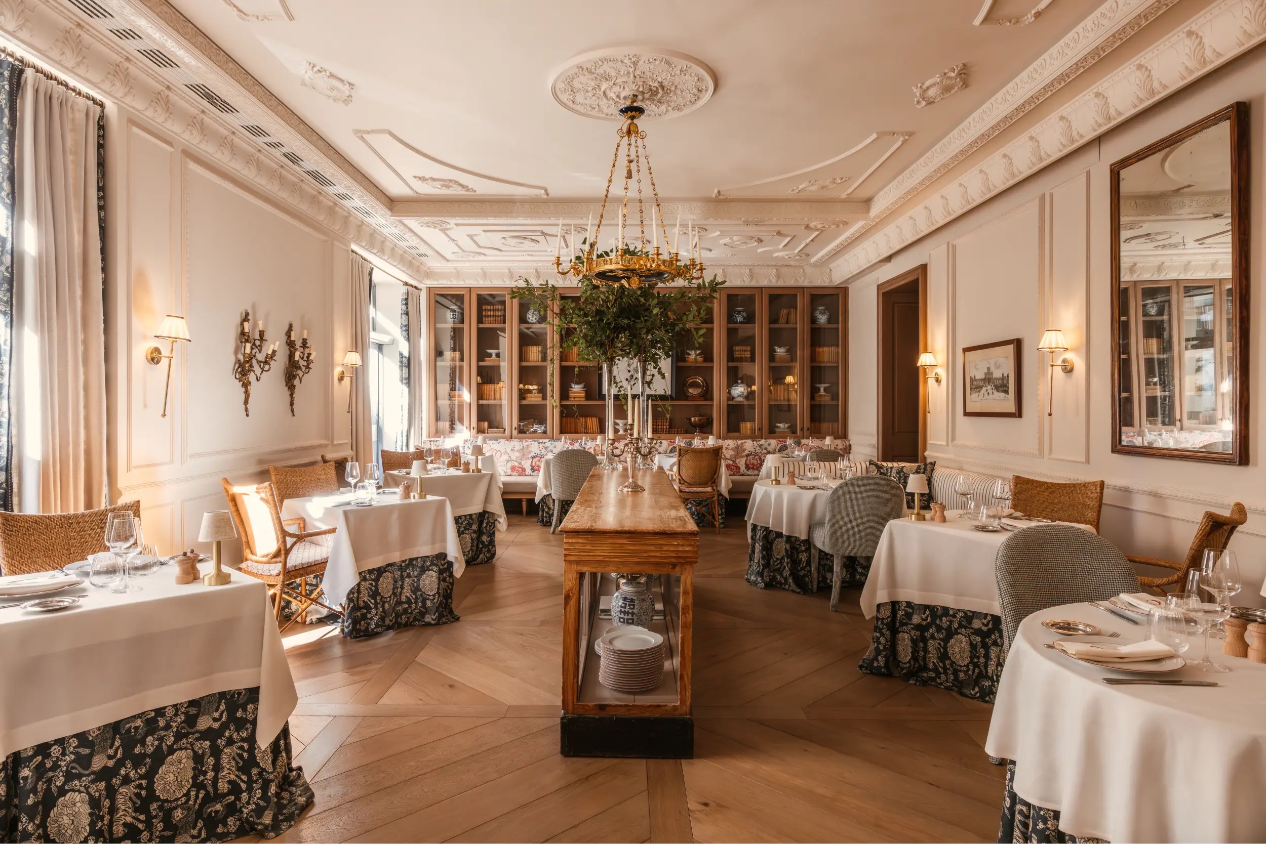A dining room with white-clothed tables over blue floral skirting, wicker and grey upholstered chairs, a gilt candle chandelier below moulded plasterwork, and glass-fronted bookcases along the back wall.