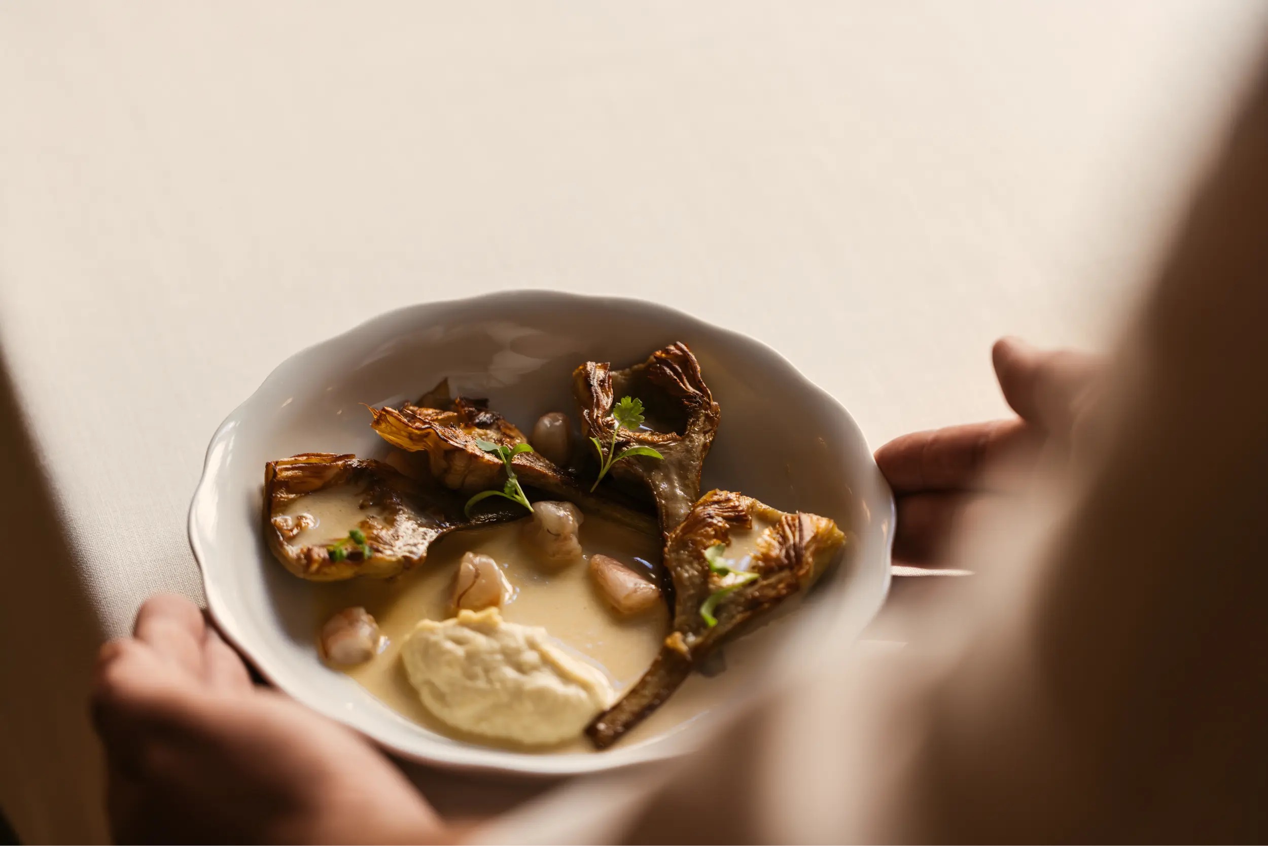 Roasted artichoke pieces with a pale puree, shellfish and herb leaves in a light sauce, served in a scalloped white bowl held over a cream tablecloth.