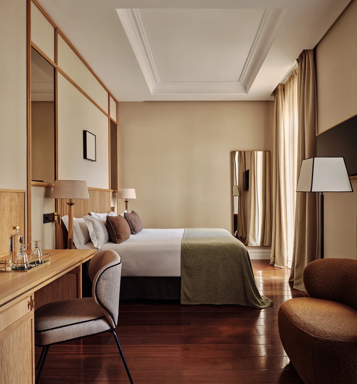 A guest room with a bed dressed in white linen and a green throw, wood-framed panelling, a wooden desk with a water tray and a corner armchair.