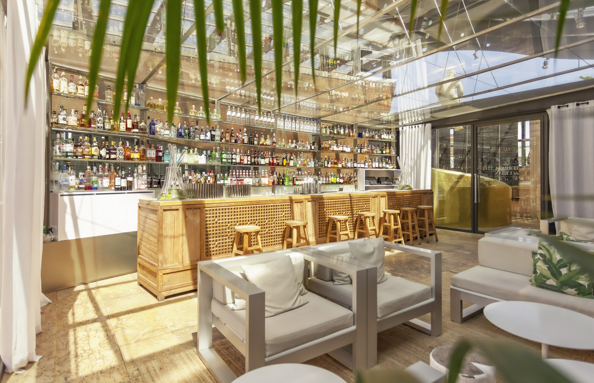 A glass-roofed rooftop bar with a wooden counter and stools, tiered mirrored shelves of bottles behind it, and pale cushioned lounge chairs and sofas in front.