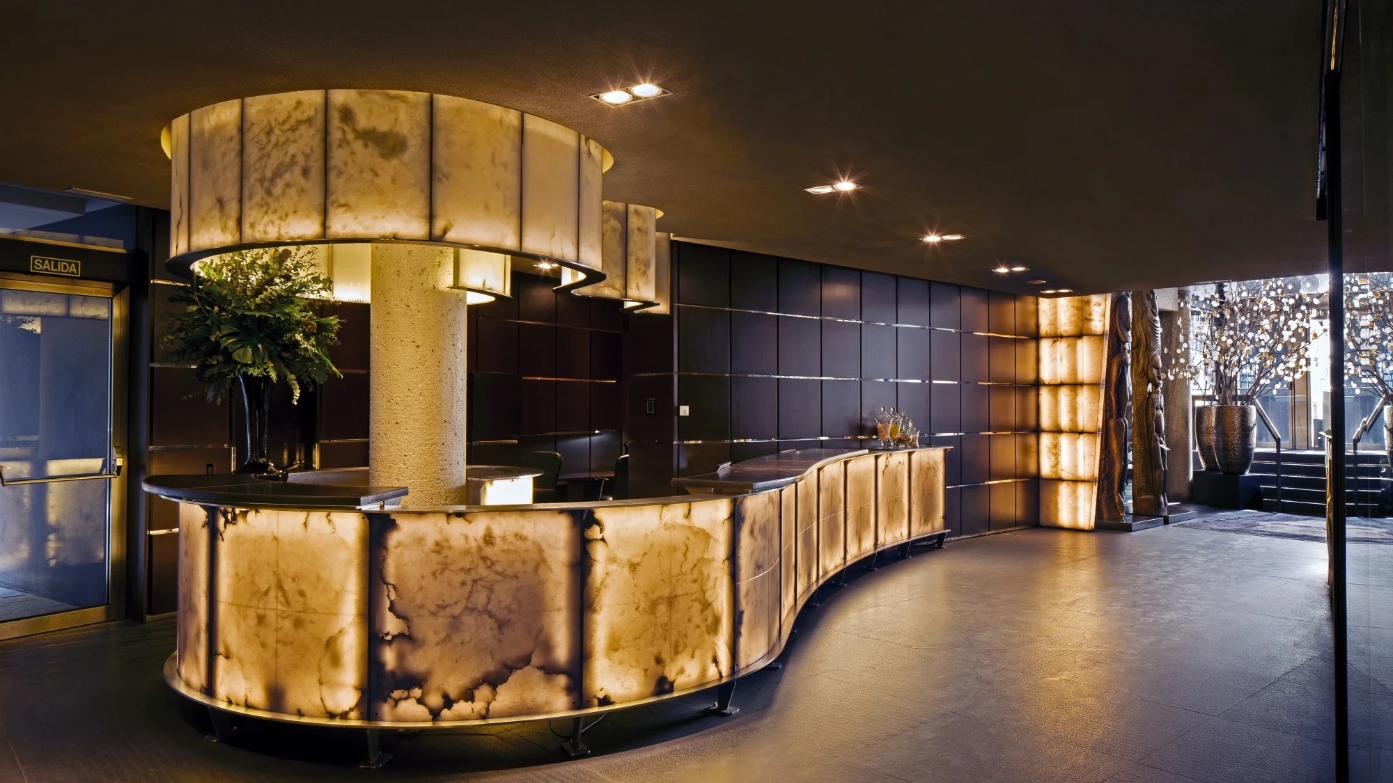 The reception hall at night: a curving desk of backlit alabaster panels around a stone column, dark panelled walls and carved wooden sculptures by the entrance.