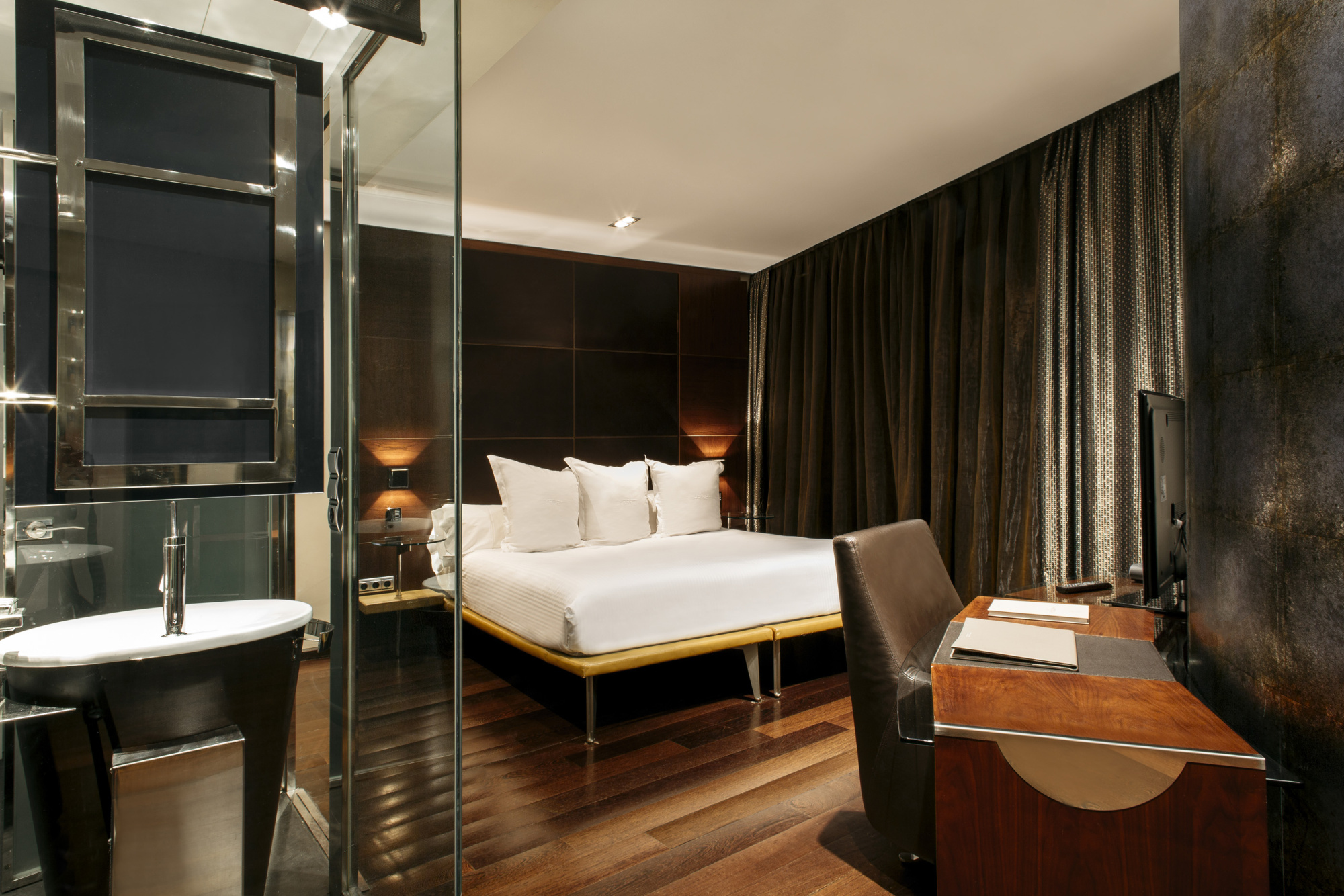 A guest room with a white-linen bed on a low platform against a dark panelled headboard wall, a wooden desk and leather chair, and a bathroom with a freestanding tub behind a glass partition.