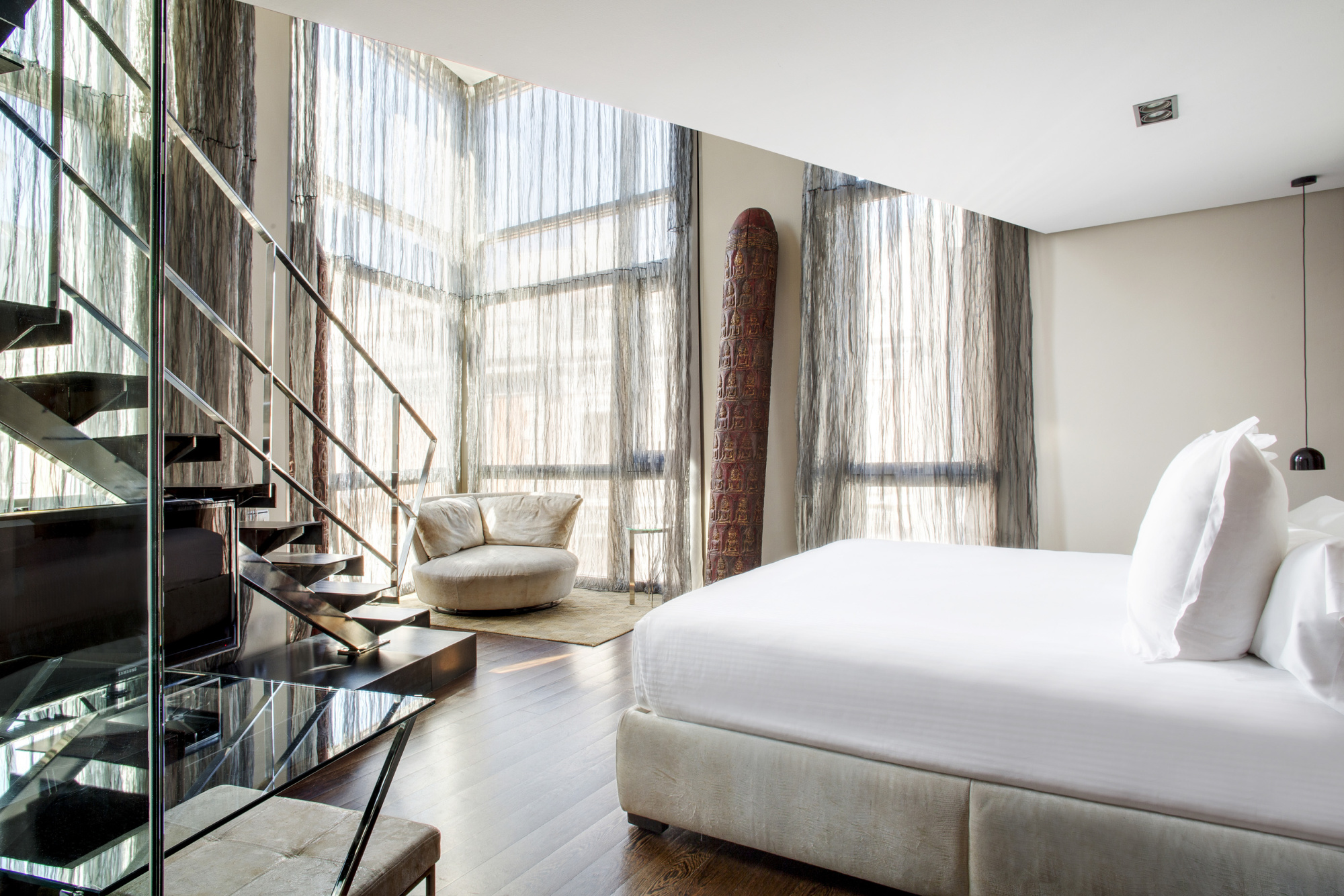 A duplex suite with a dark metal-and-glass staircase rising to an upper level, double-height windows hung with sheer curtains, a carved standing sculpture and a white-linen bed.